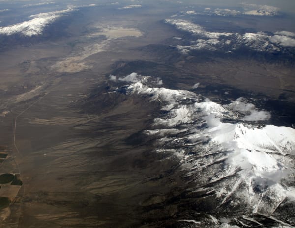 A high-altitude aerial photograph of a snow-covered desert range in the Great Basin, taken from a plane — dark forested and bare slopes streaked with snow rising to a white summit ridge under scattered cloud, a wide brown valley floor beyond it with a straight road and green centre-pivot circles at the lower left, and further snow-capped ranges receding into haze. The frame carries none of the features the park is known for: no caves, no bristlecone pines, no night sky.