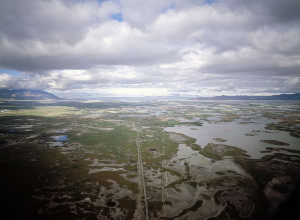 An aerial view across the marshes at the edge of the Great Salt Lake — braided shallow ponds and pale salt flats threaded by a dead-straight dike road running toward the water, with the open lake a thin bright band on the horizon and mountain ranges beyond under heavy cloud.