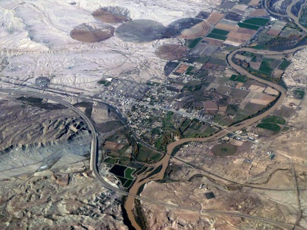 An aerial photograph looking down on a small town on a brown, meandering river, its grid of streets set among green and tan fields with circular irrigation marks. Pale eroded badlands fill the left of the frame and a highway and railway cut diagonally across it.