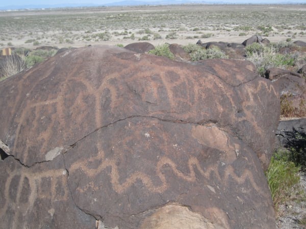 A large basalt boulder at Grimes Point covered with pecked petroglyphs — zigzags, wavy lines, and concentric and grid-like forms cut through the dark desert varnish to the paler rock beneath, the carvings continuing across a deep crack that splits the boulder in two. More dark boulders and grey-green sagebrush run back from it to the open floor of the Lahontan Valley and a hazy range on the horizon.