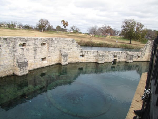 Clear spring water in a stone-walled pool at the old Hancock Springs bathhouse in Lampasas, with the park beyond.