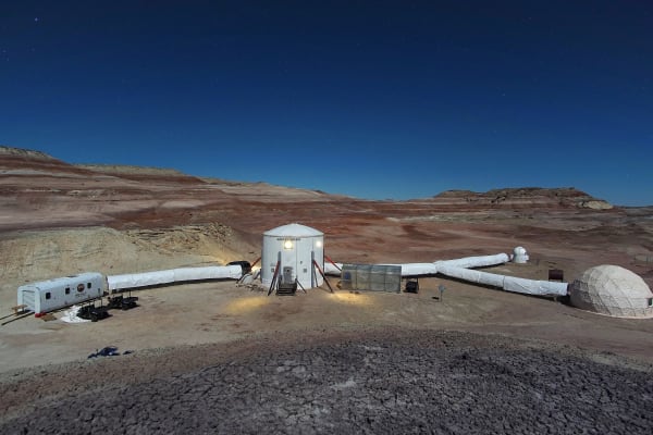 The Mars Desert Research Station at night — a white cylindrical habitat lit from outside, linked by fabric tunnels to a greenhouse, an observatory dome and a smaller dome, with a trailer and a rover parked alongside. Red and white banded badlands run back behind it under a deep blue star-flecked sky.