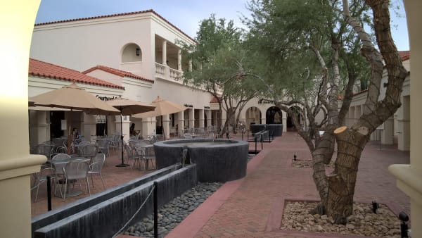 The courtyard of the Heard Museum in Phoenix seen through an arch — white stucco ranges with red tile roofs and arcaded openings down both sides, a colonnaded upper loggia with a balustrade at the far end, and shopfronts lettered Coffee Cantina and Books. Tan canvas umbrellas shade metal café tables and stacked chairs on the left, where two people are sitting; a long dark stone water channel and a round basin run down the centre over a bed of river cobbles; and mesquites with heavily pruned limbs stand in gravel beds set into the brick paving. The museum's galleries and its collections of Native art are not in the frame.