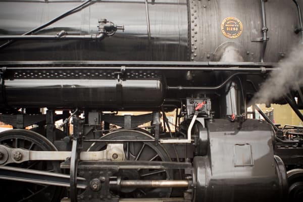 A close side view of a black steam locomotive, showing the boiler with its riveted seams, a brass builder's plate reading 31250, the driving wheels, connecting rods and air pump, with steam venting from a fitting at the right.