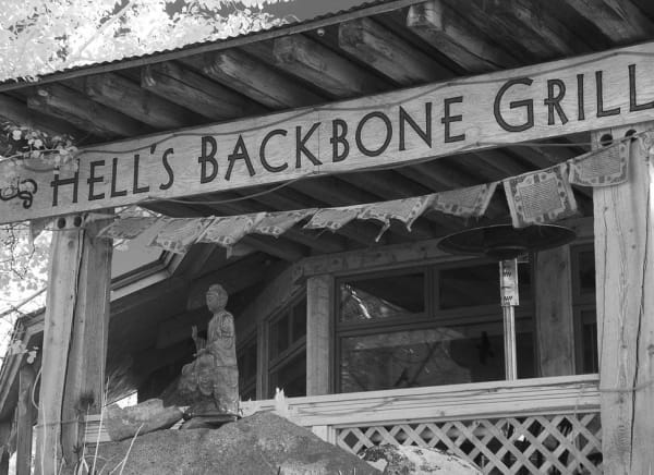 A black-and-white view of the entrance at Hells Backbone Grill, looking up at the hand-lettered timber beam spelling HELL'S BACKBONE GRILL across the front of the porch, with a string of printed prayer flags hung beneath it and a rough log post at either end. A small seated figure carved in stone stands on a boulder below the rail, with lattice screening, a patio heater and the restaurant's windows behind, and sunlit leaves crowding the frame at left.