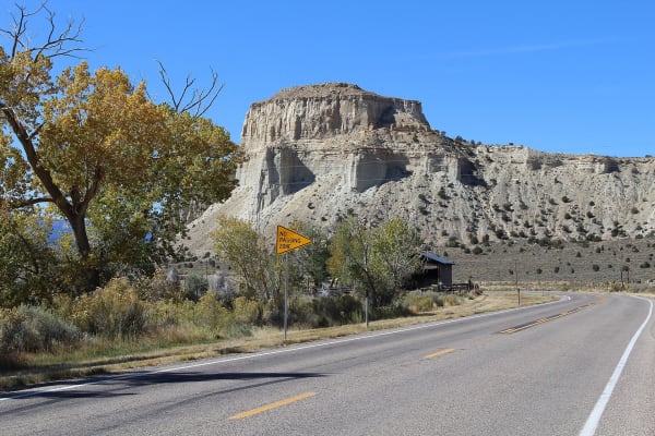 Scenic Byway 12 running past Henrieville, the empty two-lane pavement curving away at right with a yellow NO PASSING ZONE pennant on the shoulder. A cottonwood in yellow autumn leaf stands at left, a weathered timber barn with a metal roof and a pole corral sit in the middle distance, and a broad butte of pale grey slopes beneath a cream caprock rises behind, under clear deep blue sky.