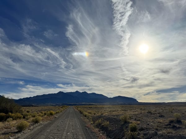 A gravel road running dead straight across sagebrush and rabbitbrush flats toward the Henry Mountains, the range standing as a long dark silhouette of rounded summits along the horizon. High cirrus fans across most of the sky, with the low sun burning white at the right and a short bar of rainbow-coloured light beside it.