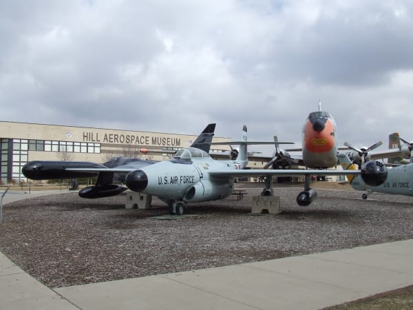 Aircraft on outdoor display at the Hill Aerospace Museum — a grey U.S. Air Force jet with wingtip fuel tanks and a black nose cone standing on a gravel apron in the foreground, propeller-driven aircraft ranged behind it including one with an orange-painted nose, and the low museum building carrying its name in raised letters across the wall beyond, under heavy grey cloud.