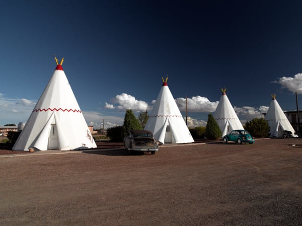 Four of the fifteen concrete teepees of Wigwam Village No. 6 at Holbrook, white cones banded with a red zigzag and topped with painted poles, each with a small door and a square window at its base, ranged around a gravel forecourt under a deep blue sky with cumulus building behind them. A rusted 1950s sedan stands between the first two and a turquoise Volkswagen Beetle beside the third, both parked as permanent exhibits. The units are a 1950 roadside imitation of a Plains teepee, marketed under the wrong word for it. No part of the town of Holbrook is in the frame.