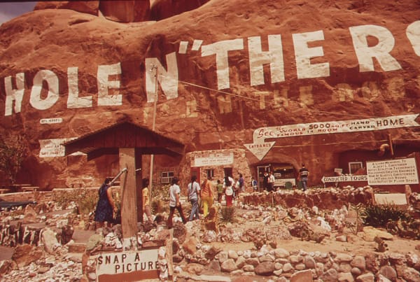 A May 1972 photograph of Hole N" the Rock south of Moab, the name painted in tall white letters across the red sandstone cliff above the carved-out house with a ghost of an earlier painting showing through beneath it, hand-lettered signs advertising conducted tours of the fourteen-room home, and visitors walking a rock-lined path through the yard.