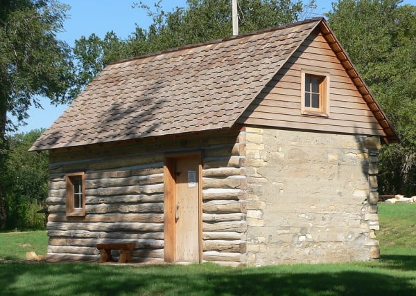 The small restored Home on the Range cabin of stone and logs, with a shingled roof, under shade trees.