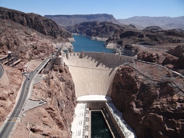 Hoover Dam curving between the cliffs of Black Canyon, Lake Mead behind it, the powerhouse wings and the Colorado River in the gorge below, and the highway switchbacking down to the crest.