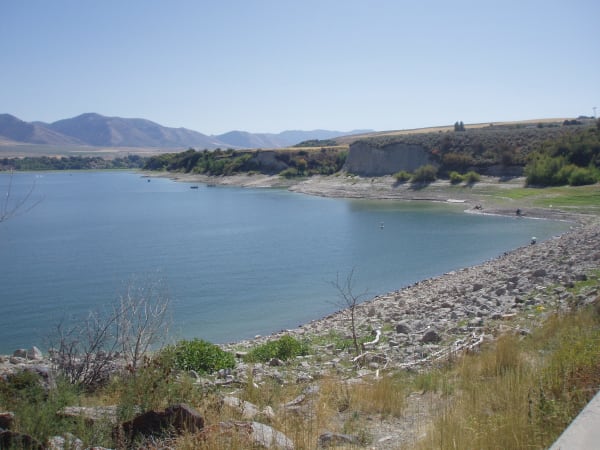 A reservoir with a stony shoreline curving away in the foreground, the water pale blue and the drawn-down bank showing bare rock and gravel. Low dry hills and a cut bluff stand along the far shore, with a few small boats out on the water.