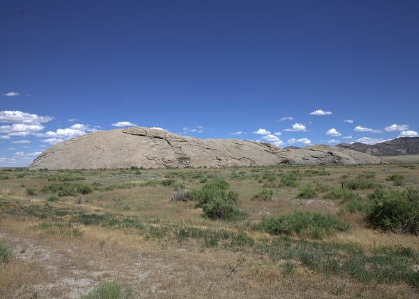 Independence Rock, a long grey granite dome rising out of sagebrush flats, seen from the west under a deep blue sky