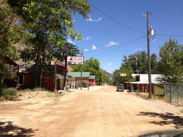 Jarbidge's dirt main street on a summer afternoon — a red board-and-batten building with an upper balcony at the left, a white-fronted storefront with a porch beside it, and a tall roadside sign reading RED DOG SALOON, FOOD BOOZE AT ITS BEST, LUNCH AND DINNER. A log building with a green metal roof stands beyond, two vehicles are parked at the roadside, and cottonwoods and canyon walls close the view.