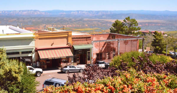 Jerome seen from above its own main street, the town built along a shelf on the side of Cleopatra Hill — a row of storefronts under striped awnings with PAUL & JERRY'S SALOON lettered across one, a roofless brick shell standing gutted and fenced beyond it, and cars parked at the kerb. An orange-berried pyracantha fills the near foreground and a ponderosa stands at the right. Beyond the roofs the ground falls away across the whole Verde Valley to red rock and a long blue escarpment on the horizon. The town is plainly occupied, and no mine workings are in the frame.