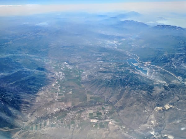 A high aerial view of a long valley, a small town strung along a straight road among a patchwork of fields, with ridges and canyons falling away on both sides and a reservoir at the right. Haze softens the distant mountains.
