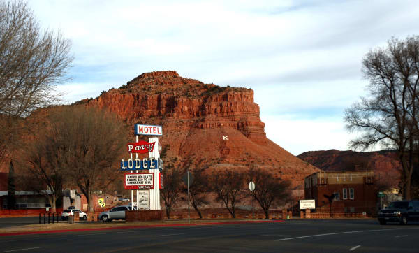 Kanab's Center Street — US-89 — on a winter afternoon, looking across wide, nearly empty pavement at the Parry Lodge sign: MOTEL in red on white, Parry in script on a red banner, LODGE in bulb-lit letters on blue, and a reader board offering HAPPY HOLIDAYS, ROOMS $59 TO 89, BREAKFAST INCL above VACANCY. Bare cottonwoods line the street, cars stand at the kerb, and a sign for the Kanab Heritage Museum fronts a brick building at right. A red sandstone butte rises behind the town in late light, a white letter K on its slope.