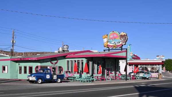 Mr. D'z Route 66 Diner at Kingman — a long single-storey building painted mint green with magenta trim, its rooftop sign carrying a burger, fries and a turquoise cartoon car above ROUTE 66 DINER lettering. A blue 1950s pickup with Route 66 shields on its door is parked at the kerb beside a pink novelty microcar, turquoise and magenta picnic tables and furled red umbrellas stand along the frontage, and a gift shop with its own Route 66 signs adjoins to the right. A dark basalt bluff closes the view at the left and the wide asphalt of the highway crosses the foreground. The railroad the town grew around is not in the frame.