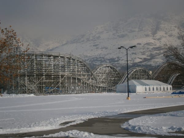 Lagoon's wooden roller coaster standing empty in winter, its white timber trestle running in a long series of humps across the frame, seen across a snow-covered lot with a marquee tent and a lamp standard beside it and the snow-dusted Wasatch foothills rising into haze behind.