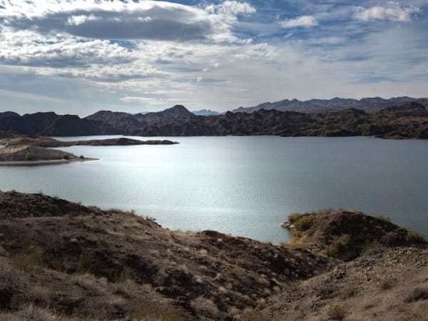 A wide bay of Lake Mead seen from a bare hillside above it, brown desert ridges enclosing the water on every side, a low green-tufted spit reaching out from the near shore and a pale band of bare rock above the waterline.