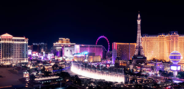 The Las Vegas Strip at night from above, a fountain display lit white across the water in the foreground and the boulevard beyond it massed with neon — an illuminated observation wheel, a half-scale Eiffel Tower and rows of lit hotel towers running to the dark desert horizon.