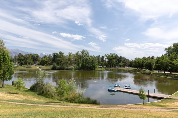 The boating pond at Liberty Park in summer — three blue pedal boats with canopies moored along a low wooden dock, a wooded island standing in the middle of the green water, and mature trees ringing the far bank. Mown lawn slopes down in the foreground, and the snow-flecked peaks of the Wasatch show above the treeline at left.