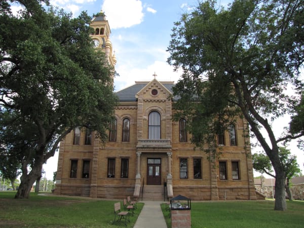 The tan brick Llano County Courthouse with its clock tower, seen up a walk between large oaks on the courthouse square.
