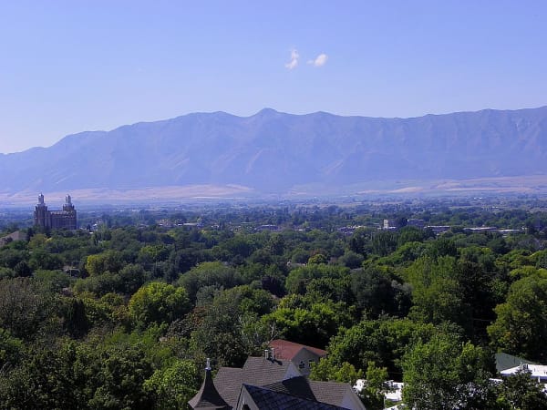 A valley town seen from a hillside above it, dense summer tree cover hiding most of the streets, with a pale stone temple and its towers standing clear at the left. Beyond the rooftops the valley floor runs flat to a long mountain wall under a hazy blue sky.
