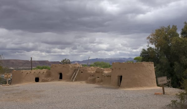 Reconstructed Ancestral Puebloan rooms on the grounds of the Lost City Museum — low mud-plastered walls with dark doorways and a pole ladder leaning against one roofline, an interpretive sign at the right, under heavy grey cloud with mesas on the far horizon.