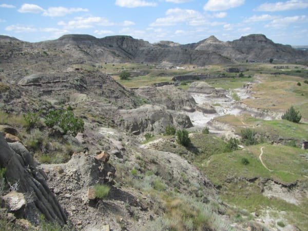 Eroded grey and buff badlands at Makoshika, a pale wash winding through green draws below a line of buttes