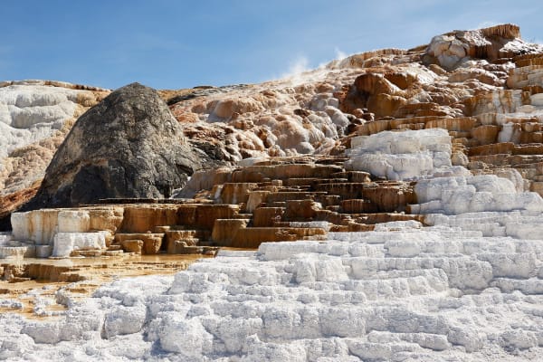 White and orange travertine terraces stepping down a slope at Mammoth Hot Springs, a dark boulder among them and steam rising behind