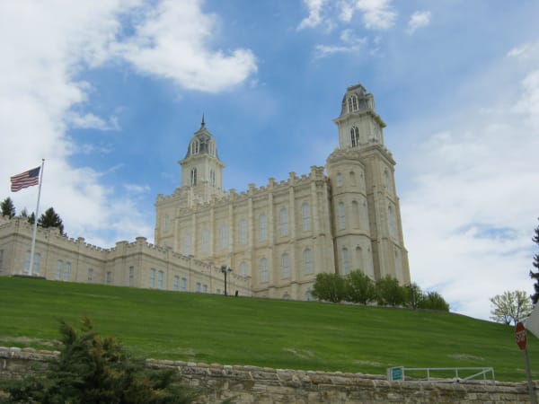 The temple seen from the foot of its hill, looking steeply upward: cream oolite limestone walls beneath a castellated parapet, rows of tall arched windows, and two towers, the taller one at the right. A mown green slope and a rough stone retaining wall fill the lower half of the frame.