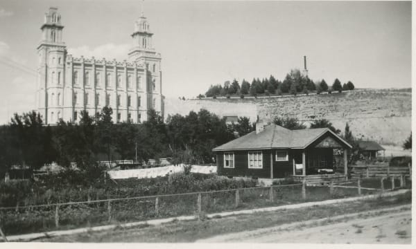 An archival black and white photograph looking across a dirt road and fenced garden plots toward the temple standing on its hill. Laundry hangs on lines, a small bungalow with a porch sits at the right, and a bare quarried hillside topped with conifers and a tall chimney rises beyond.