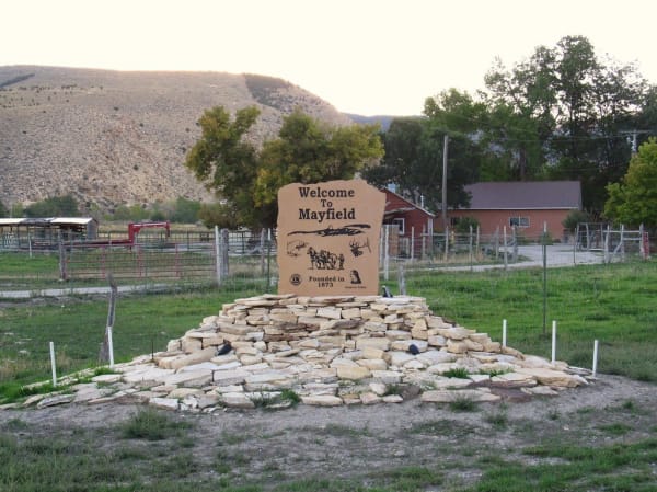 A carved sandstone welcome monument on a low mound of stacked flagstones, reading Welcome To Mayfield above a ridgeline, a trout, a two-horse team with a teamster, an elk head, and Founded in 1873. A carved profile of a Native man sits at the lower right beside the words Arapeen Valley, with a corral, a red barn and a dry hillside behind.