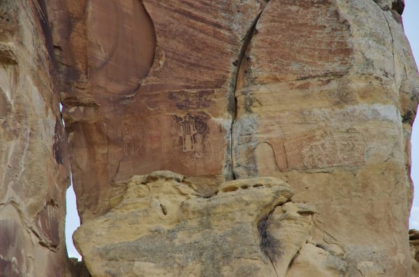 A Fremont-era petroglyph panel on a sandstone cliff in Dry Fork Canyon — broad-shouldered, trapezoidal human figures pecked pale through the dark red desert varnish, several wearing headdresses and necklaces, with round patterned discs beside and below them. The panel sits partway up the wall, pale gold ledges below it and weathered red-brown rock above.