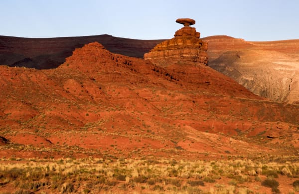 Mexican Hat Rock in low evening sun — a broad flat disc of sandstone roughly sixty feet across, balanced on a narrow neck at the top of a small butte and reading exactly as a sombrero seen edge-on. The butte beneath it is banded orange and red in horizontal ledges and falls away into a long slope of red siltstone streaked with shadow and dotted with low brush. Dry pale grass fills the foreground, and banded grey and tan cliffs stand across the background in shadow.