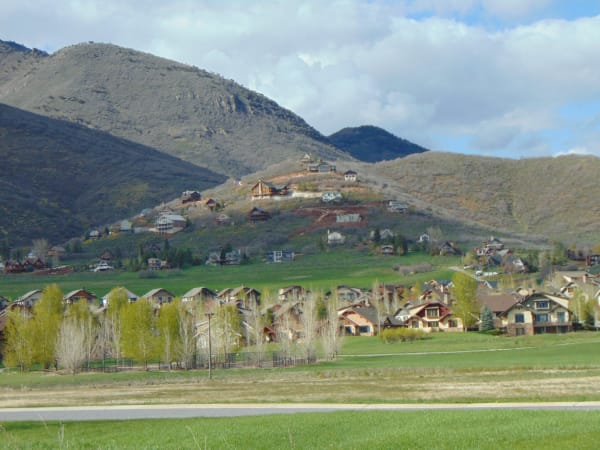 Modern chalet-style houses with steep gables spread across a green valley floor and up onto the sagebrush hillside behind, a mown field and a paved path in the foreground and a bare rounded peak rising under broken cloud.