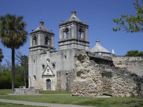 The twin bell towers and dome of Mission Concepcion in San Antonio, with ruined stone walls of the convento in front and a palm at left.