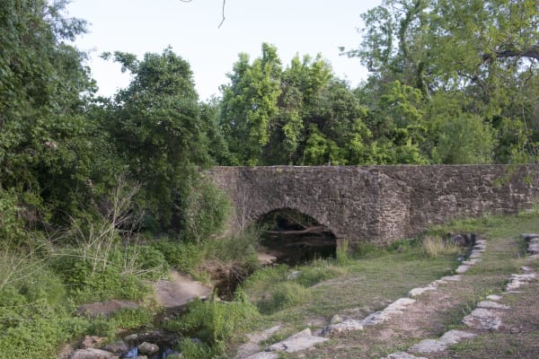 The two stone arches of the Espada Aqueduct carrying the acequia over Sixmile Creek in San Antonio, among green trees.
