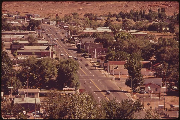 An archival photograph looking down the town's main street from above, a wide highway lined with gas station signs for Standard and Conoco, low storefronts, a motel sign and parked cars of the period. Cottonwoods fill the side streets and bare red hills rise behind the town.