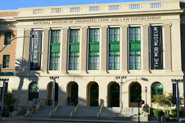 A pale neoclassical civic building with arched ground-floor openings and green-framed windows, NATIONAL MUSEUM OF ORGANIZED CRIME AND LAW ENFORCEMENT lettered across the frieze and THE MOB MUSEUM on tall banners hung either side of the entrance steps.