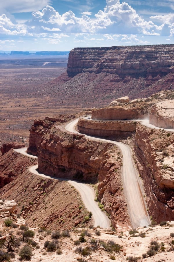 The Moki Dugway seen from high on the cliff — three tiers of unpaved switchback cut into the face of Cedar Mesa, the pale gravel road doubling back on itself across ledges of red sandstone with a low stone and earth berm along the outer edge and no guardrail anywhere on it. A light pickup runs one of the upper bends trailing a long plume of dust. Past the last turn the road straightens out across the tan desert floor eleven hundred feet below, a broad cliff-walled mesa stands to the right of it, and flat-topped buttes line the far horizon under high summer cumulus.