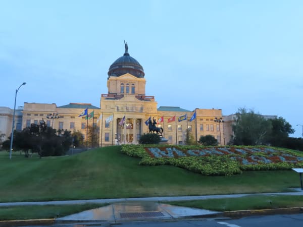 The Montana State Capitol in Helena, sandstone wings and a copper dome lit at dusk above a sloping lawn and flower bed