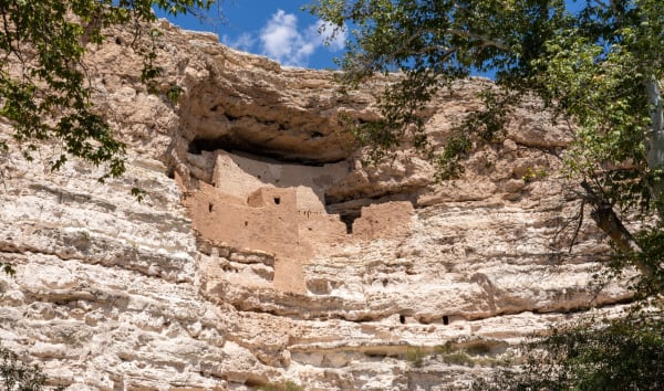 Montezuma Castle from directly below, a block of mud-plastered masonry standing several storeys high in a deep undercut in a pale limestone cliff, its rooms stepped back under the overhang with small dark door and window openings across the front. Bands of eroded limestone run the width of the frame above and below it, sycamore branches hang in at both edges, and a strip of blue sky with small cumulus shows over the rim. It was built by the people archaeologists call Sinagua; the name on the map is not theirs, and no emperor was ever here.