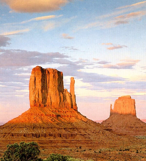 A scanned printed reproduction of the Mittens in Monument Valley at low sun. West Mitten Butte stands at the left, a broad red sandstone tower with a slender detached spire beside it, rising from a talus skirt above the stepped ledges of the valley floor; East Mitten Butte stands smaller and further off at the right with its own thin spire. Warm cloud streaks a pale blue and pink sky, and juniper and sage cover the foreground bench. The frame carries the fine dot screen of the printing it was scanned from.