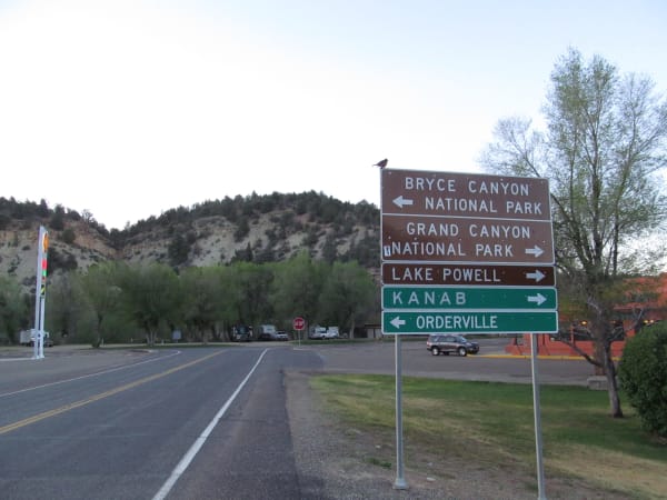 The junction signpost at Mount Carmel — a stack of five direction signs reading BRYCE CANYON NATIONAL PARK with a left arrow, then GRAND CANYON NATIONAL PARK, LAKE POWELL and KANAB with right arrows and ORDERVILLE with a left, with a small bird perched on the top edge. The empty highway runs past in the foreground, a stop sign and low motel buildings stand at the far corner, and a wooded sandstone bluff rises behind.
