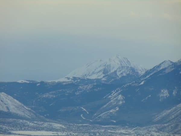 A snow covered peak with a sharply serrated summit ridge seen at a distance through winter haze, above layered blue grey foothills. A town spreads across the pale valley floor along the bottom of the frame and the sky above is a flat wash of haze.
