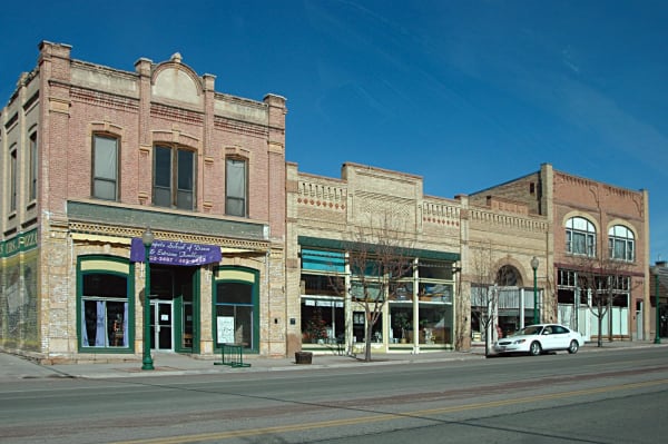 A block of 19th-century brick commercial buildings on Mount Pleasant's Main Street seen obliquely down the street, arched upper windows and patterned brick cornices above shopfronts with green-framed glass, BANK lettered into one parapet, a faded painted ghost sign at the left edge, bare street trees and green lamp standards along the sidewalk and a single car at the kerb.