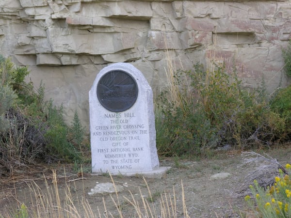 A white stone marker with a bronze medallion naming Names Hill and the old Green River crossing, set before the pale sandstone cliff where emigrants carved their names